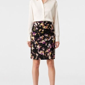 May include: A woman wearing a cream-colored button-down shirt and a black pencil skirt with a floral pattern in shades of purple, yellow, and white. She is also wearing black high heels.