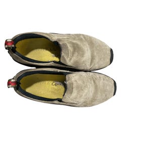 May include: Pair of beige slip-on shoes with black soles and trim. The shoes have a worn appearance, with yellow insoles and a red, white, and black pull tab on the heel. The shoes are designed for casual wear.