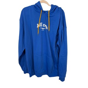 Puede incluir: Una sudadera con capucha azul con un bolsillo delantero y una capucha con cordones amarillos. La sudadera con capucha tiene texto blanco que dice "kill crew" y otro texto debajo. La sudadera con capucha está colgada en una percha de madera.