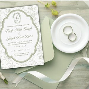May include: A wedding invitation card featuring a sage green ornate frame, floral background patterns, and the text Emily Anne Cassidy and Joseph Scott Ricker. The set includes a sage green envelope, two silver rings, and a white ribbon.