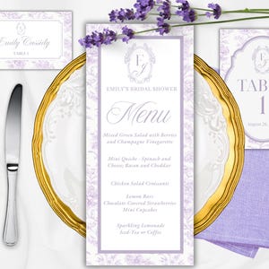 May include: A bridal shower stationery set featuring a menu card, place card, and table number card with a lavender floral pattern, gold-rimmed plate, silver fork and knife, and a purple napkin. The menu reads Emily's Bridal Shower Menu.