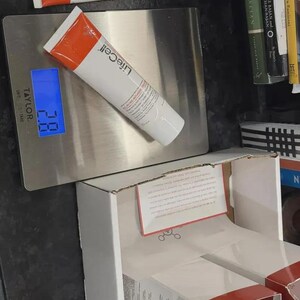K&ouml;nnte beinhalten: Wei&szlig;e und orangefarbene Tuben von LifeCell South Beach Skincare liegen auf einer silbernen Waage, die 2,8 anzeigt. Daneben befindet sich eine wei&szlig;e Schachtel mit roten Akzenten und einem bedruckten Einsatz.