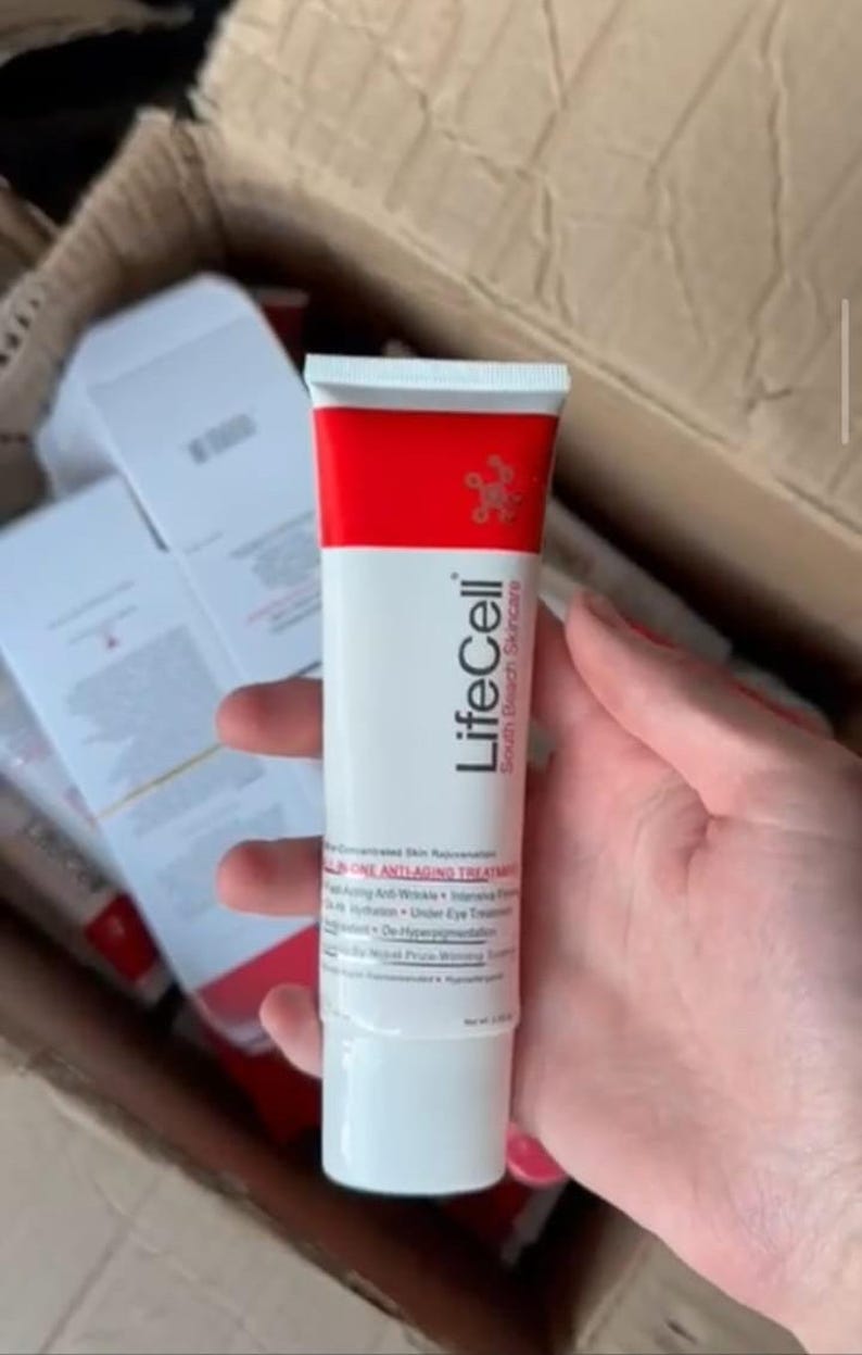 K&ouml;nnte beinhalten: Eine wei&szlig;e und rote Tube LifeCell South Beach Skincare ONE ANTI-AGING TREATMENT wird gehalten. Die Tube hat einen roten Deckel und einen wei&szlig;en K&ouml;rper mit schwarzem Text. Weitere Kartons sind im Hintergrund zu sehen.