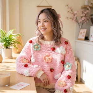 May include: A light pink crochet sweater adorned with smiling flower and strawberry appliqués, along with white cloud accents. The sweater has a round neckline and ribbed cuffs. The overall design is whimsical and handmade.