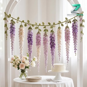Op de afbeelding: Een gehaakte slinger met hangende wisteria-achtige bloemen in tinten paars, lavendel en crème, geaccentueerd met groene bladeren. De slinger is gedrapeerd over een witte boog. De tekst ZENVYCO DIGITAL is zichtbaar in de rechterbovenhoek.