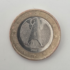 Puede incluir: Una moneda de un euro con un detallado emblema de águila plateada en el centro, rodeado por un anillo dorado. El borde de la moneda presenta un patrón de estrellas y el año 2002 es visible. La moneda mide aproximadamente 2,3 cm de diámetro.