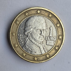 May include: A one Euro coin featuring a portrait of a man, possibly a historical figure, with the text "1 EURO" and the year "2002". The coin has a gold-colored outer ring with stars.