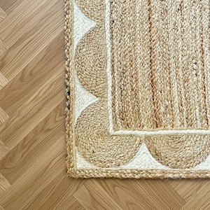 Pode incluir: Um tapete retangular feito de fibras de juta natural de cor castanha, com um painel central tecido rodeado por uma borda recortada branca. O design incorpora uma textura entrançada, com a borda recortada a criar um padrão semicircular repetitivo ao longo do perímetro do tapete.