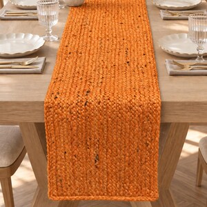 Può includere: Un runner da tavola rettangolare realizzato in fibre intrecciate di colore arancione con sottili macchie marrone scuro. Il runner presenta una spessa trama intrecciata ed è centrato lungo un tavolo da pranzo in legno chiaro.