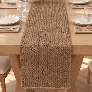 Puede incluir: Un camino de mesa rectangular hecho de fibras de yute trenzadas en color marrón natural con un tejido grueso y texturizado. El camino está centrado en una mesa de comedor de madera clara, flanqueado por platos blancos festoneados, cubiertos dorados y cristalería estriada.
