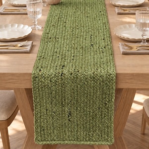 May include: A rectangular table runner crafted from braided green natural fibres, featuring a thick, textured weave with visible variations in the green tone. The runner is positioned lengthwise down the centre of a wooden dining table.