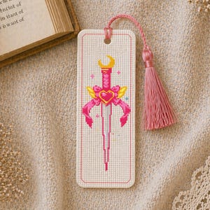 May include: A handmade bookmark with a cross-stitch design of a pink sword with a heart-shaped hilt, yellow accents, and a crescent moon. The bookmark has a pink tassel attached to the top. The bookmark is rectangular with a white background.