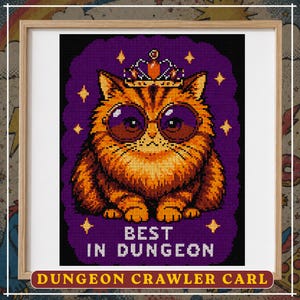 May include: A framed cross-stitch pattern featuring an orange tabby cat wearing a crown and round purple sunglasses. The cat is set against a dark purple background with yellow stars and the white text BEST IN DUNGEON below the cat.