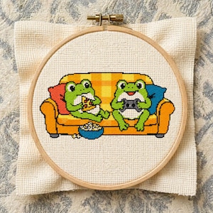 May include: Cross-stitch embroidery in a wooden hoop. The design features two green frogs on a yellow couch, one eating pizza and the other playing a video game. A bowl of popcorn sits in front of them.