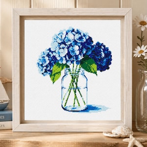 May include: Cross-stitch art of blue hydrangea flowers in a clear glass jar. The flowers have varying shades of blue and green leaves. The artwork is framed in a light wood frame and sits on a wooden surface.