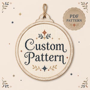 Może przedstawiać: Okrągły, kremowy wzór haftu krzyżykowego w drewnianej ramie ze słowami "Custom Pattern" w kolorze granatowym. Rama ma jutowy sznurek do zawieszenia. Mała etykieta głosi "PDF PATTERN". Zawiera dekoracyjne akcenty kwiatowe i gwiazdki.