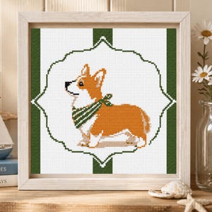 Könnte beinhalten: Kreuzstich-Kunstwerk eines Corgi-Hundes mit einem grün-weiß gestreiften Halstuch. Der Corgi ist orange und weiß, vor einem weißen Hintergrund in einem grün-weiß gestreiften Rahmen. Das Kunstwerk befindet sich in einem hellen Holzrahmen.