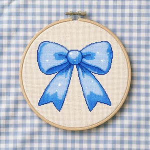 Può includere: Ricamo a punto croce di un fiocco blu in un telaio di legno. Il fiocco presenta una sfumatura di blu chiaro e scuro, con riflessi bianchi. Lo sfondo è un tessuto chiaro con un motivo a quadretti blu e bianchi.