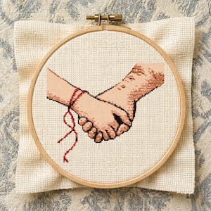 May include: Cross-stitch embroidery of two hands clasped together, with a red string tied around one wrist. The design is set within a wooden embroidery hoop and rests on a cream-colored fabric.