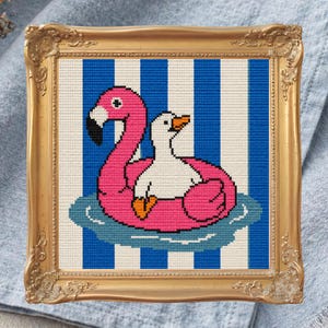 Puede incluir: Obra de arte de punto de cruz en un marco dorado. La imagen presenta un flotador de flamenco rosa con un pato blanco encima, sobre un fondo a rayas azules y blancas. La obra está enmarcada y lista para colgar.