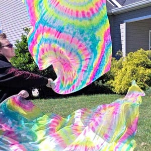 May include: Brightly colored tie-dye fabric in shades of pink, yellow, blue, and green is held up in the air. The fabric is flowing and appears to be lightweight. The background includes a house and green grass.