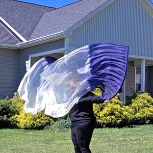 May include: A flowing, two-toned flag, white and deep purple, is held aloft against a bright blue sky. The flag is being waved in front of a house with a green lawn and bushes. The flag's colors create a dynamic visual effect.