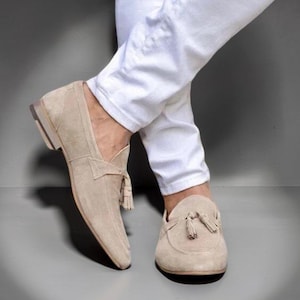 Peut inclure: Une paire de mocassins en daim beige avec un petit talon et un détail à pompons sur le dessus de chaque chaussure. Les mocassins sont portés avec un pantalon blanc, présentant un bout pointu et une finition mate sur la surface extérieure.