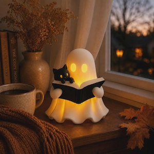 May include: A white ghost-shaped lamp with glowing yellow eyes, holding an open black book. A small black cat figure sits on the top edge of the book. The lamp emits a warm light from within its smooth, rounded, translucent body structure.