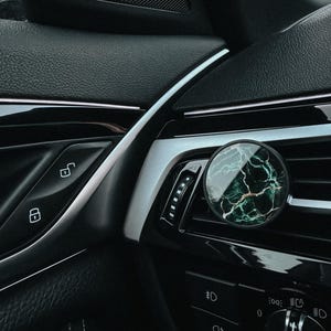 Könnte beinhalten: Ein Auto-Luftauslass-Clip mit einem runden, marmorähnlichen Design in Grün und Weiß. Der Clip ist an einem schwarzen Armaturenbrett mit silbernen Akzenten befestigt. Der Innenraum des Autos hat schwarzes Leder und glänzende schwarze Zierteile.