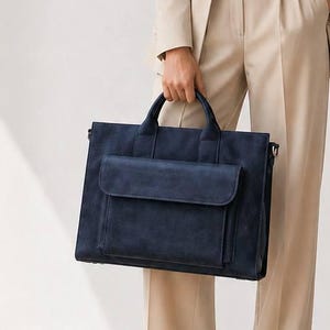 Op de afbeelding: Een vrouw in een beige pak en zwarte hakken houdt een marineblauwe suède aktetas vast. Het pak bestaat uit een blazer en een broek met wijde pijpen. Ze draagt een witte blouse, een bril en een ketting. De achtergrond is een effen witte muur.