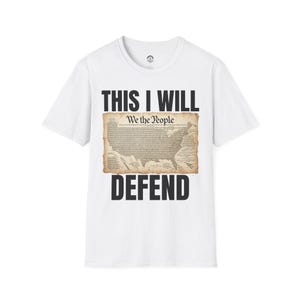 May include: White t-shirt with the text "THIS I WILL DEFEND" in black block letters. The shirt features a vintage-style image of the United States map with the words "We the People" printed above it.