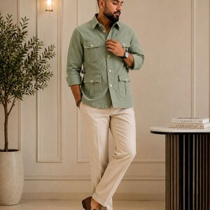 May include: A sage green long-sleeve button-down shirt featuring four front flap pockets and side waist adjusters, paired with beige trousers and brown leather loafers. The shirt has a classic collar and light-colored buttons down the front.