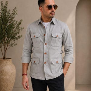 Pode incluir: Uma camisa de manga comprida cinza-claro com botões, quatro bolsos frontais com aba, botões castanhos e tiras de ajuste laterais na cintura. Usada sobre uma camisola branca, combinada com calças pretas e óculos de sol com armação dourada.