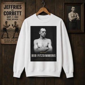 May include: A white long-sleeve sweatshirt featuring a black and white photographic print of boxer Bob Fitzsimmons with his arms crossed. The text reads "Ruby Robert", "BOB FITZSIMMONS", "THREE DIVISION CHAMPION", "CORNWALL", and "1890s".