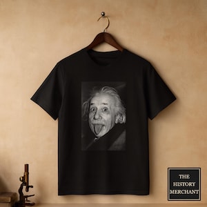 Op de afbeelding: Een zwart t-shirt met korte mouwen en een gecentreerde zwart-wit fotoprint van Albert Einstein die zijn tong uitsteekt. Het shirt hangt aan een houten kledinghanger tegen een neutrale achtergrond, met een klein logo in de hoek.
