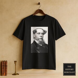 Pode incluir: Uma t-shirt preta de manga curta com uma impressão gráfica retangular centrada de um retrato a preto e branco de Charles Dickens. O retrato mostra um homem com uma barba cheia e cabelo distinto, vestindo um casaco escuro e camisa branca.