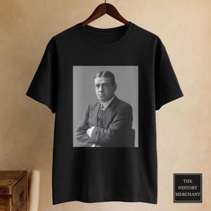 Könnte beinhalten: Ein schwarzes T-Shirt mit kurzen Ärmeln und einem mittig platzierten, quadratischen Schwarz-Weiß-Fotodruck eines Mannes im Anzug mit verschränkten Armen. Das T-Shirt hängt an einem Holzkleiderbügel. Ein kleines schwarzes Quadrat mit dem Text THE HISTORY MERCHANT ist zu sehen.