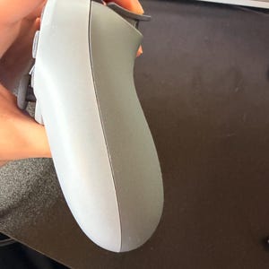 Könnte beinhalten: Ein grau-dunkelgrauer Videospiel-Controller mit Tasten und Triggern. Der Controller wird in einer Hand gehalten, mit einer Tastatur und einer dunklen Oberfläche im Hintergrund.