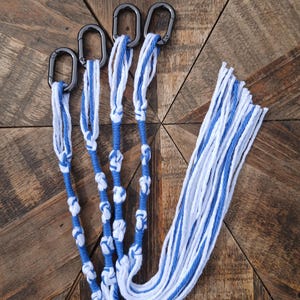 Peut inclure: Suspension pour plante en macramé avec cordon bleu et blanc. Le support comporte quatre mousquetons en métal noir en haut, avec des détails noués le long des cordons bleus et blancs. Les extrémités des cordons sont effilochées.