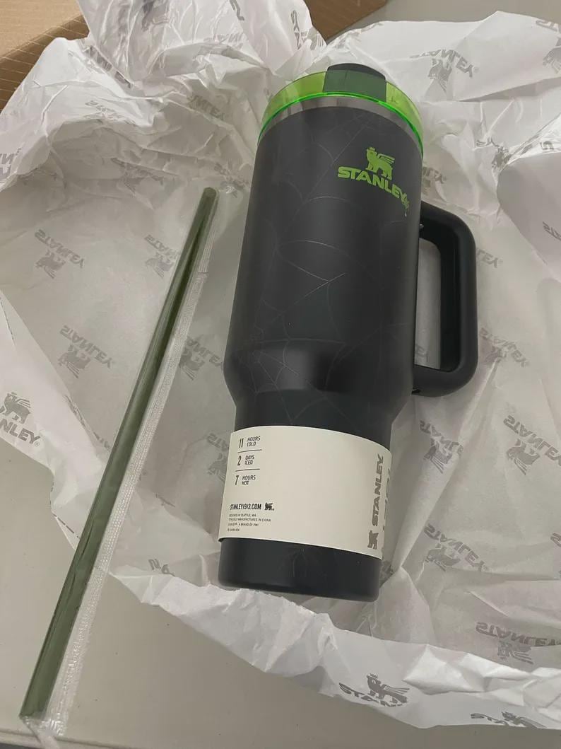 May include: A black Stanley mug with a green lid and handle, and a green straw. The mug has the Stanley logo in green and a white label. The mug is sitting in white tissue paper.