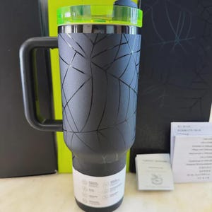 May include: A black insulated tumbler with a handle and a green lid. The tumbler has a geometric design etched into the surface. The tumbler is designed to keep beverages hot or cold. The tumbler is shown with a black box and some paperwork.