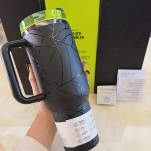 May include: Black insulated tumbler with a geometric design and a handle. The tumbler has a neon green lid and a black handle. The tumbler is made of stainless steel and is designed to keep drinks cold or hot. The tumbler is approximately 9 inches tall.