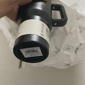 May include: Black Stanley Quencher Tumbler with a handle. The tumbler has a white label with the Stanley logo and product information. The handle is black and attached to the side of the tumbler. The top of the tumbler is green.