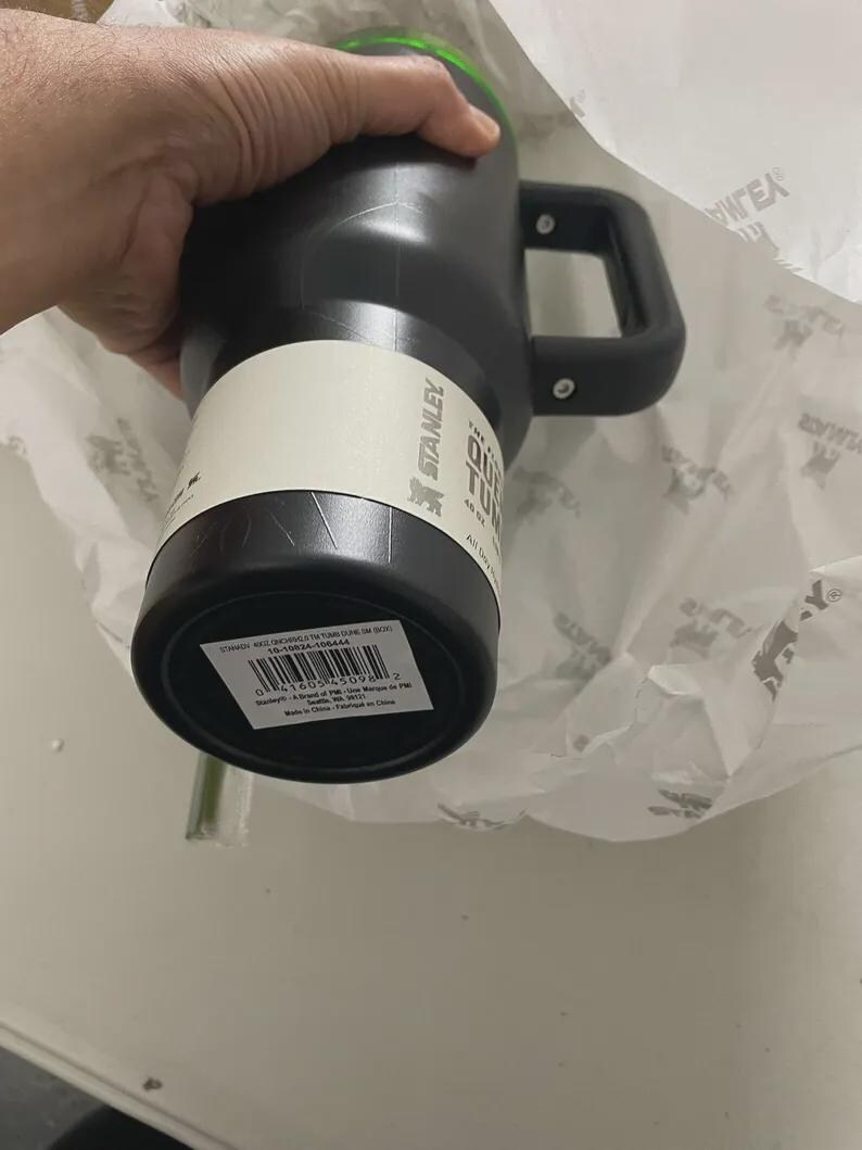May include: Black Stanley Quencher Tumbler with a handle. The tumbler has a white label with the Stanley logo and product information. The handle is black and attached to the side of the tumbler. The top of the tumbler is green.