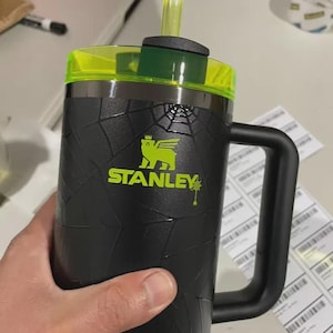 May include: Black Stanley mug with a neon green lid and straw. The mug has a textured design and a large handle. The Stanley logo is in neon green. The mug is held by a person.