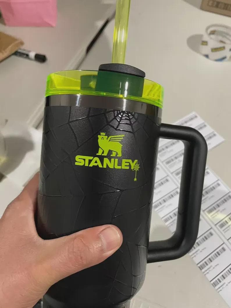 May include: Black Stanley mug with a neon green lid and straw. The mug has a textured design and a large handle. The Stanley logo is in neon green. The mug is held by a person.