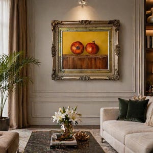 May include: A framed painting with an ornate gold frame shows two red pomegranates on a wooden surface against a yellow background. The artwork is displayed on a neutral wall in a living room, with a sofa and decorative items.