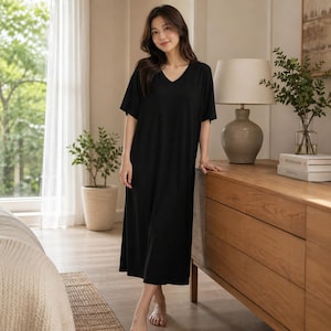 May include: A black, short-sleeved V-neck dress. The dress is floor-length and made of a soft, flowing fabric. The model is standing in a well-lit room with a window and a wooden dresser. The dress is a comfortable and stylish choice for lounging or casual wear.
