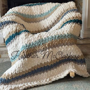 Peut inclure: Une couverture en tricot épais à rayures crème, bleues et beiges. La couverture est drapée sur une chaise, mettant en valeur sa texture douce et sa palette de couleurs. La couverture porte le texte "Heather McCloskey" en écriture cursive.