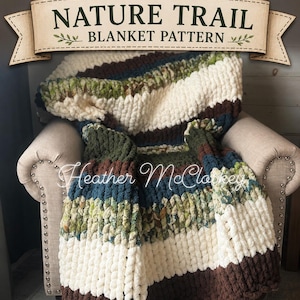 Op de afbeelding: Een dikke gebreide deken in bruin, crème, groen en blauw, gedrapeerd over een beige fauteuil. De deken heeft de tekst "Nature Trail Blanket Pattern" en de naam "Heather McCloskey".